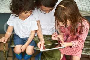 Engaged in a whole wide world online. three adorable little children using a digital tablet while sitting on a bench outdoors.