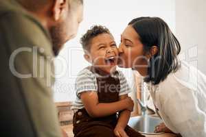 Happy boy getting a kiss by caring mother, bonding and laughing during family time at home. Young parents sharing a sweet moment of parenthood with their playful child, relaxing and carefree together
