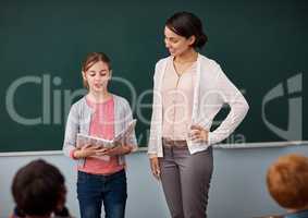 Shes a confident speaker. an elementary school girl reading in front of the class while standing alongside her teacher.