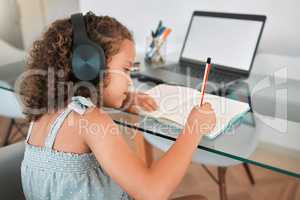 Homeschooling by a smart and clever little girl attending online or virtual class using a laptop and headphones. A young child writing and working or doing homework at home listening to music