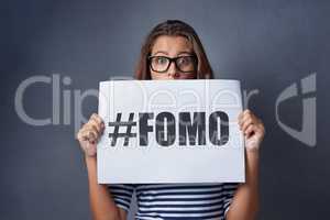 If it happened online it must be real. Studio shot of an attractive young woman holding a sign with FOMO printed on it against a gray background.