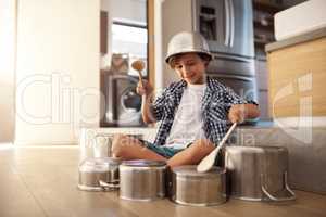 Hes a little noisemaker. a happy little boy playing drums with pots on the kitchen floor while wearing a bowl on his head.