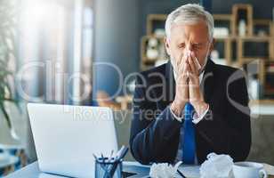 I cant afford to be sick now. a frustrated businessman using a tissue to sneeze in while being seated in the office.