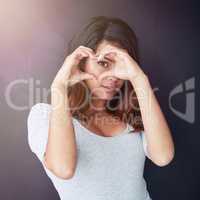 Seeing everything through love. Portrait of a beautiful young woman looking through her heart-shaped hands in the studio.