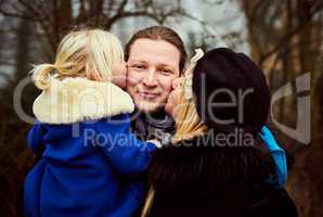 Kisses from my two favourite girls. a mother and daughter kissing the father on the cheeks outdoors.