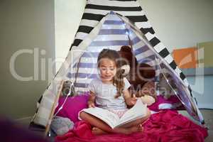 Books can help fuel a childs curiosity and imagination. a little girl reading a book with her teddybear in a tent at home.