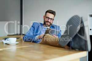 Business and leisure done in one. a young businessman working on a digital tablet with his feet up on the desk in an office.