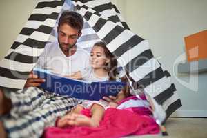 They love looking at all the pictures. a father reading a book with his little daughter in a tent at home.