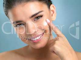 We all need pampering. Close up studio shot of a beautiful young woman applying moisturiser on her face against a blue background.
