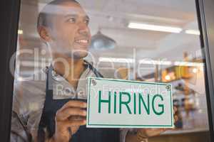 Recruitment, advertising and small business owner with hiring sign board on window or door of cafe or restaurant, happy and positive. Young business man excited open after end of covid virus pandemic